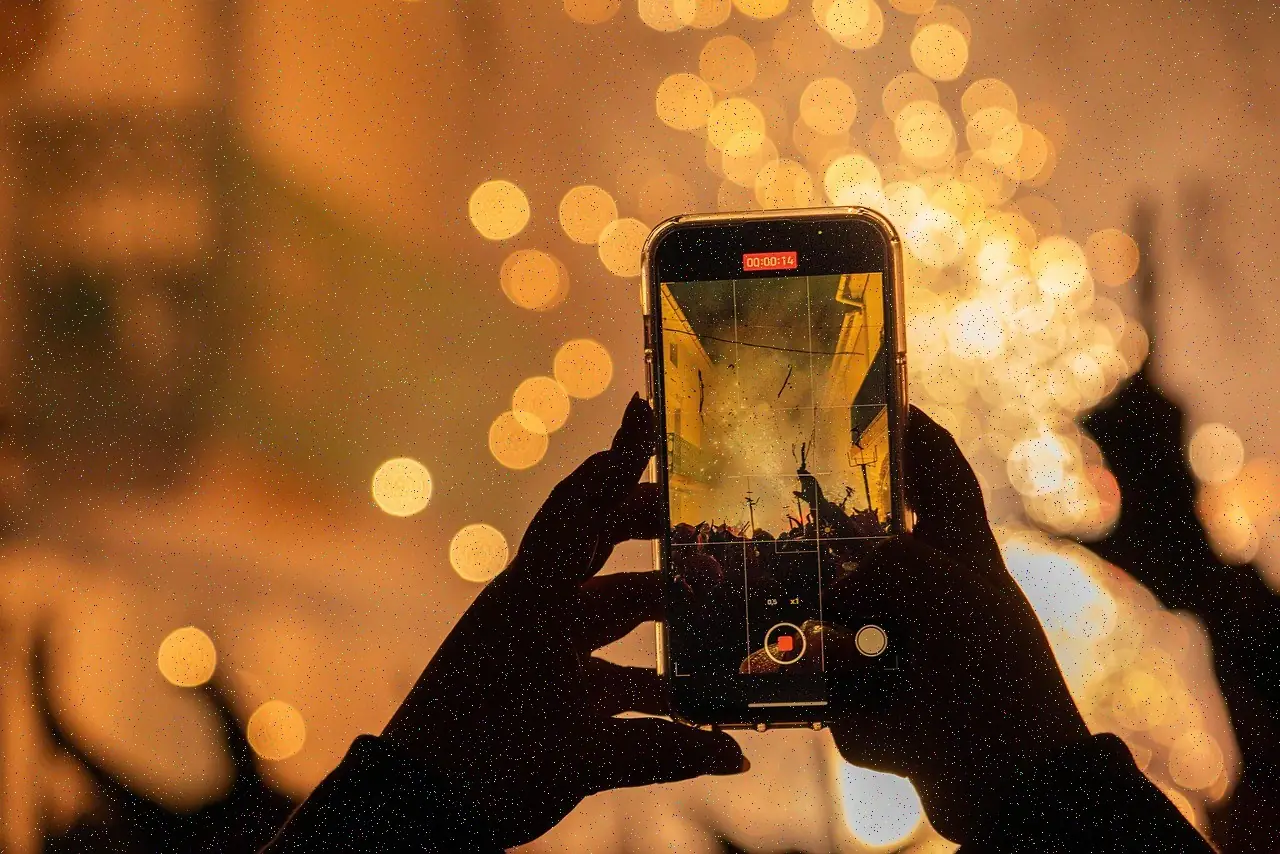 A concertgoer holds up a phone to film the stage through warm, blurred lights