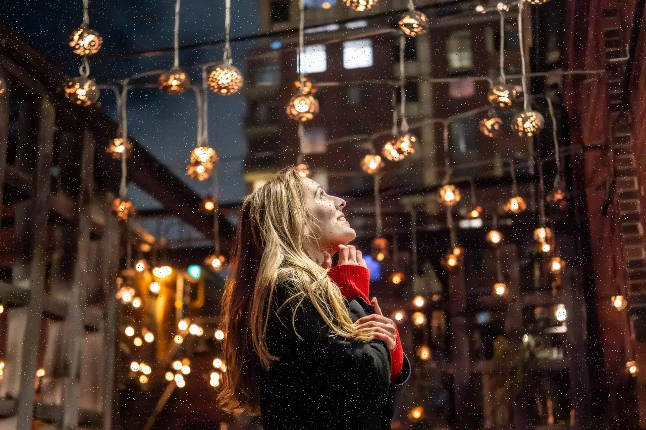 A woman looks up at strings of glowing bulbs in an evening courtyard